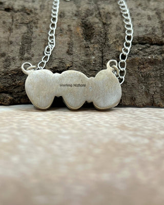 A silver heart-shaped pendant necklace with three connected hearts, engraved with 'Sterling Nizhoni', resting on a textured brown bark surface with a blurred beige foreground.