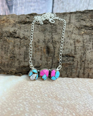 A silver chain necklace with three oval turquoise and pink stone pendants, displayed on a piece of textured wood over a light beige surface.
