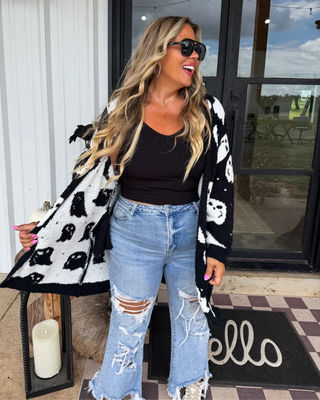 Woman wearing a black and white patterned cardigan, black top, and ripped jeans standing on a porch.