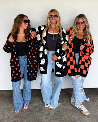 Three women wearing colorful cardigans and jeans standing against a white wall.