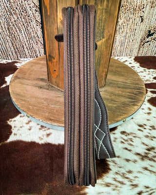 A dark brown leather zipper case or sleeve with black zippers and white contrast stitching, standing upright on a cowhide rug in front of a rustic wooden spool.