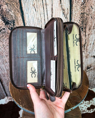 A dark brown leather wallet with multiple compartments, shown open and held by a hand over a rustic wooden surface. The wallet features card slots, a zippered pocket, and interior pockets with a light beige printed lining.