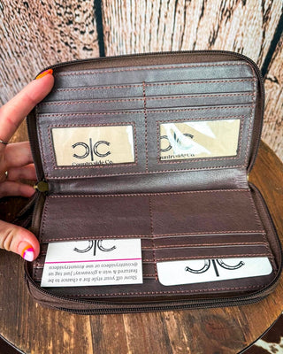 An open dark brown faux-leather wallet held by a hand, showing interior card slots, two branded nameplates reading 'Countryside Co', and a small card insert on a wooden surface.