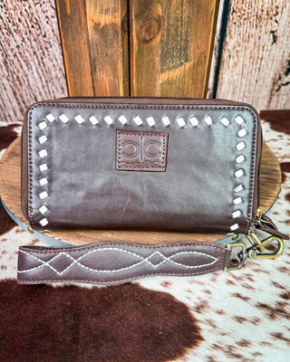 A dark brown leather wristlet wallet with silver-tone studs and a matching wrist strap, displayed on a wooden stand over a cowhide rug.