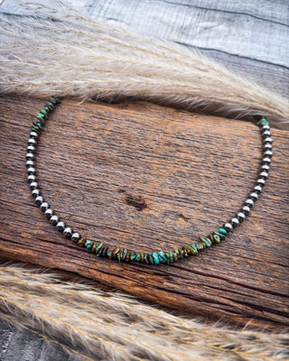 A beaded necklace with small metallic silver beads and a central cluster of irregular turquoise and brown stone beads, displayed on a rustic wooden surface with dried grass.