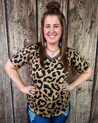 A woman wearing a tan and black leopard print short-sleeve V-neck top with a black lace-trimmed neckline, posing in front of a rustic wood plank wall.