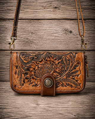 A brown tooled leather crossbody bag with floral and sunflower designs, featuring a snap-button closure and a long shoulder strap, displayed on a rustic wooden plank background.