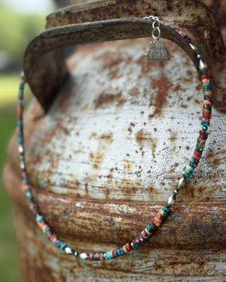 A beaded necklace with small multicolored round beads and a silver-toned charm engraved with 'Junk City', draped over a rustic rusty metal container outdoors.