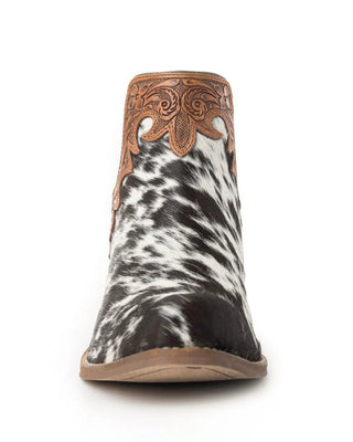 A single women's western ankle boot shown from the front on a white background. The boot features a black and white cowhide patterned lower section, a brown leather shaft with ornate cutout western embroidery, and a light brown sole.