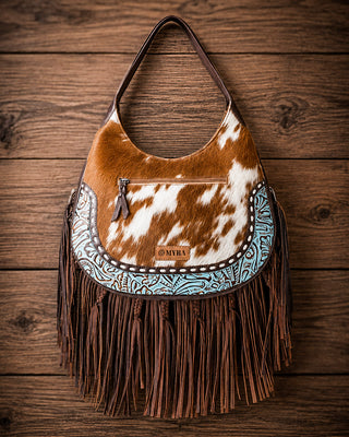 A western-style shoulder bag with a brown and white cowhide body, turquoise and brown embroidered trim, and long brown fringe, displayed against a rustic wooden plank background. A small leather patch on the front reads MYRA.