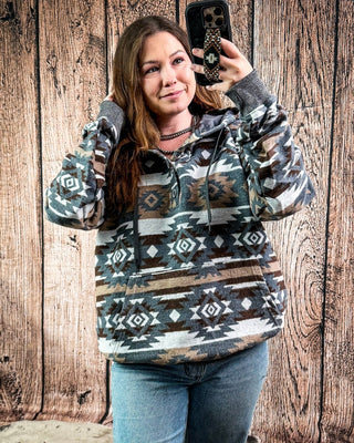 A woman takes a mirror selfie wearing a long-sleeve hooded sweatshirt with a Southwestern geometric pattern in brown, beige, gray, and white, paired with blue jeans, set against a rustic wooden plank wall.