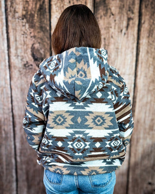 A person viewed from behind wearing a hooded sweatshirt with a Southwestern geometric pattern in slate blue, brown, beige, and white, paired with blue jeans against a rustic wooden plank background.