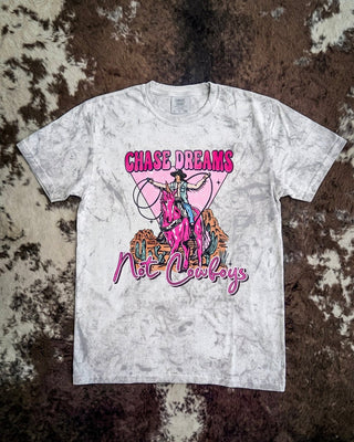 A white acid-wash graphic t-shirt featuring a cowboy illustration and the text 'CHASE DREAMS Not Cowboys' in pink, laid flat on a brown and white cowhide rug.