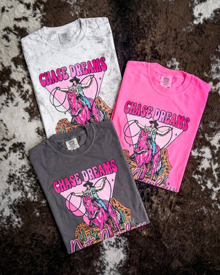 Three folded graphic T-shirts displayed on a brown and white cowhide rug. The shirts are in white, dark gray, and bright pink, each featuring a colorful graphic of a cowboy riding a horse with the text 'CHASE DREAMS' 