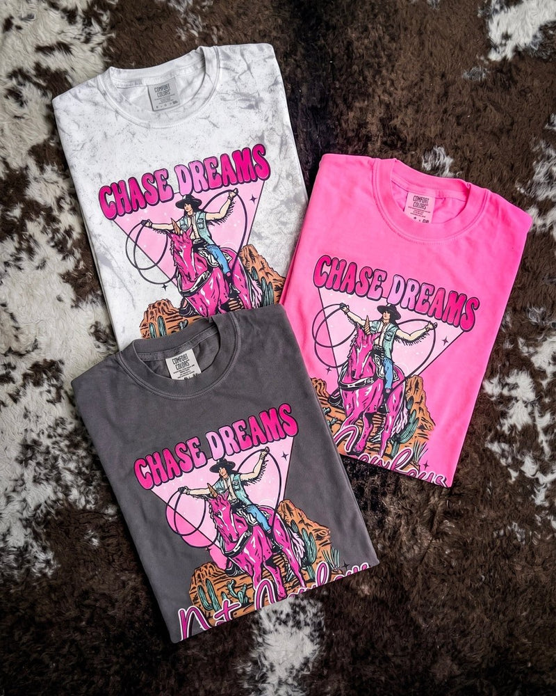 Three folded graphic T-shirts displayed on a brown and white cowhide rug. The shirts are in white, dark gray, and bright pink, each featuring a colorful graphic of a cowboy riding a horse with the text 'CHASE DREAMS' 