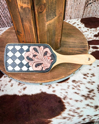 A hand-tooled leather hair brush with a black and white checkerboard pattern and pink floral detailing, resting on a rustic wooden stand over a cowhide rug.