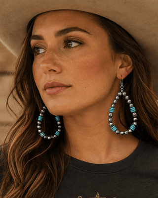A woman wearing a beige cowboy hat and a dark graphic t-shirt with a longhorn skull design, featuring large turquoise and silver beaded teardrop earrings. She is posed against a rustic wooden wall.