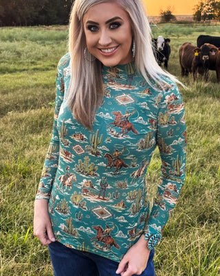 A woman with blonde hair stands in a grassy field at sunset wearing a teal long-sleeve mock-neck shirt with a detailed western-themed print featuring cacti, horses, and Native American motifs, paired with blue jeans.