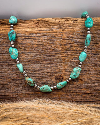 A turquoise and silver beaded necklace displayed on a rustic wooden surface with dried grass in the foreground.