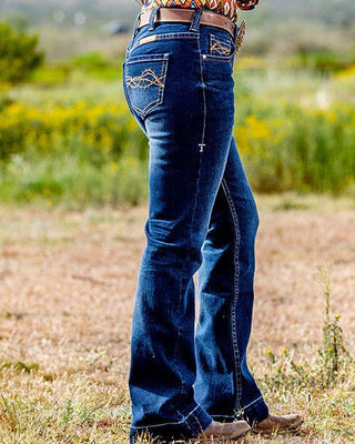 Woman wearing a colorful patterned top and blue jeans with a cowboy hat in a natural setting.