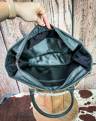 A black leather handbag shown open, with a hand holding the top edge to display the interior. The bag has a black satin-like lining with multiple pockets and is displayed on a wooden stand against a rustic wood plank background.