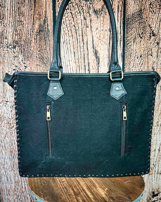 A black leather tote bag with two vertical zippered pockets and double top handles, displayed on a rustic wooden surface against a wood plank background.