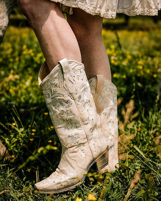 A close-up of a person's legs wearing distressed off-white western cowboy boots with floral embroidery, standing in a grassy field with yellow wildflowers.
