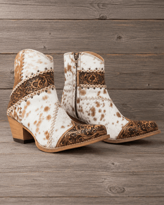 A pair of women's western ankle boots with a white and brown cowhide pattern, brown tooled leather detailing, and stacked wooden block heels, displayed on a rustic gray wood plank background.