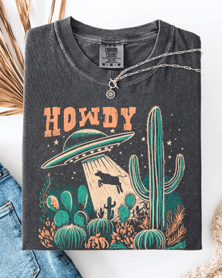 Dark gray t-shirt with 'HOWDY' and UFO design, placed on a white surface with jeans and decorative items.
