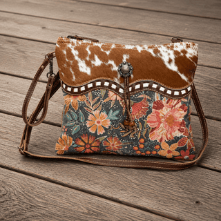 A western-style crossbody bag with a brown and white cowhide-patterned top flap, a decorative scalloped trim, and a colorful floral print body, displayed on a rustic wooden plank surface.