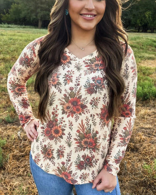 A woman wearing a long-sleeve V-neck top with a red, orange, and brown floral pattern on a cream background, paired with blue jeans, standing outdoors in a grassy field.