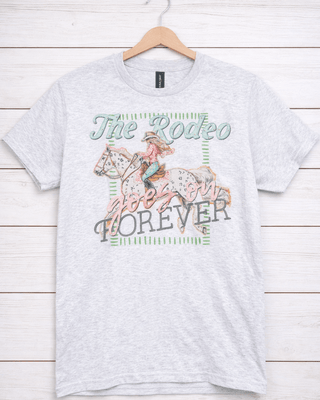 Gray t-shirt with a graphic of a person riding a horse and 'The Rodeo Goes on Forever' text on a wooden background