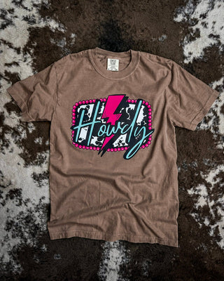 A brown short-sleeve t-shirt with a graphic reading 'Howdy' in script over a pink lightning bolt and cow print background, laid flat on a brown and white cowhide rug.