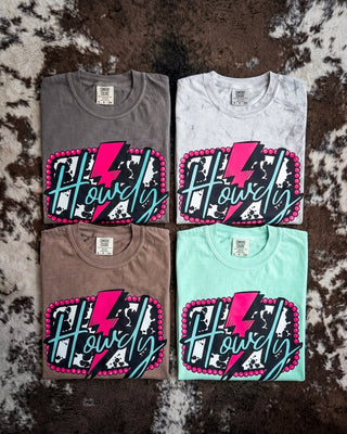 Four folded graphic t-shirts arranged in a 2x2 grid on a brown and white cowhide rug. The shirts are in dark gray, white, brown, and mint green, each featuring a large graphic on the front with a pink lightning bolt, black and white splatter pattern, and the word 'Howdy' in teal script.
