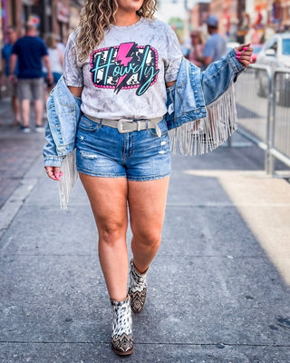 A woman walking on a city sidewalk wearing a light gray graphic t-shirt with a pink and teal 'Howdy' graphic, distressed denim shorts, a fringed light blue denim jacket draped off her shoulders, and brown and white patterned cowboy boots.