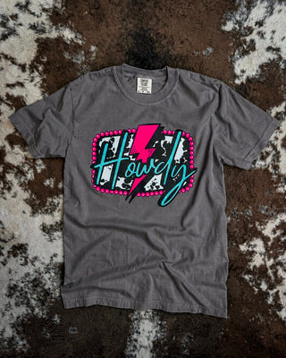 A pepper short-sleeve t-shirt with a graphic reading 'Howdy' in script over a pink lightning bolt and cow print background, laid flat on a brown and white cowhide rug.