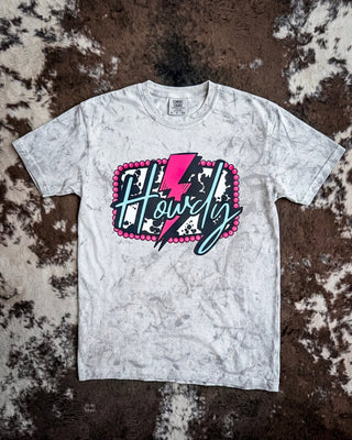 A smoke colorblast short-sleeve t-shirt with a graphic reading 'Howdy' in script over a pink lightning bolt and cow print background, laid flat on a brown and white cowhide rug.