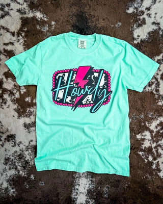 A mint green short-sleeve t-shirt with a graphic reading 'Howdy' in script over a pink lightning bolt and cow print background, laid flat on a brown and white cowhide rug.
