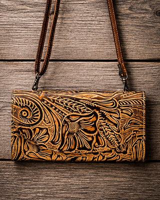 A brown tooled leather clutch bag with an intricate floral and scroll pattern, featuring a matching leather shoulder strap, displayed flat on a rustic wooden plank background.