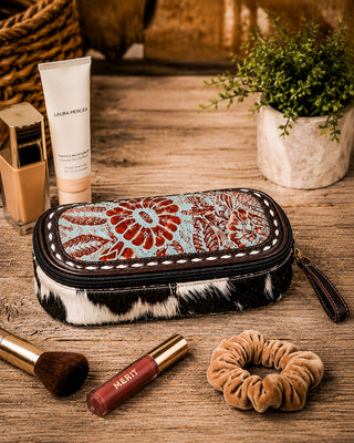 A rectangular zippered cosmetic pouch with a turquoise and red floral pattern on the front and a black-and-white cowhide-textured bottom, styled on a rustic wooden table with makeup accessories and a small potted plant.