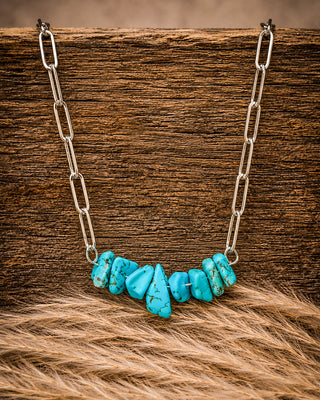 A necklace with a curved row of polished turquoise-colored stone chips on a silver paperclip chain, displayed on a rustic wooden surface with tan fur in the foreground.