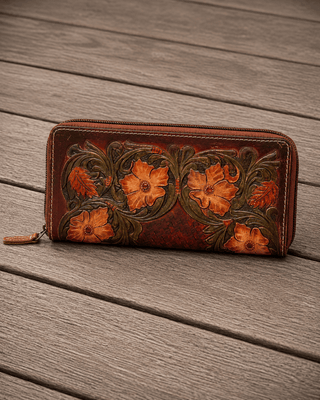 A brown leather zip-around wallet with hand-tooled floral designs in orange and green, resting on a gray wooden plank surface.