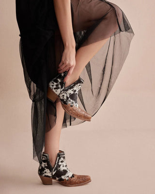 Person wearing a black dress with sheer overlay and cow print boots on a beige background