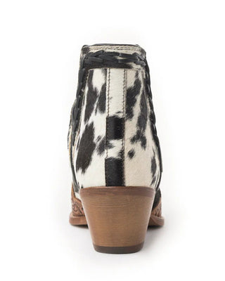 A single women's ankle boot shown from the back on a white background, featuring a black and white cow-print upper with a brown stacked block heel.