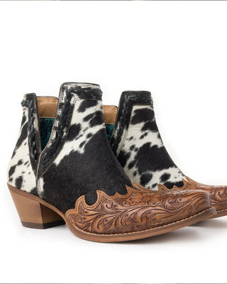 A pair of women's western ankle boots featuring a black and white cowhide pattern on the upper and a hand-tooled brown leather toe cap with floral scrollwork, set against a plain white background.