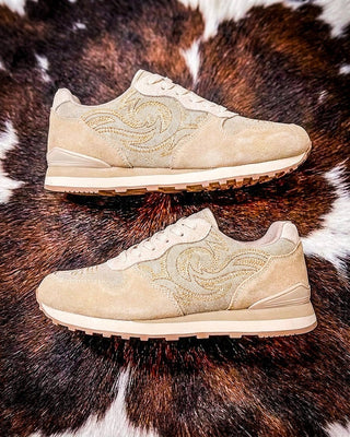 A pair of beige suede sneakers with tonal embroidered swirl patterns, displayed on a brown and white cowhide rug.