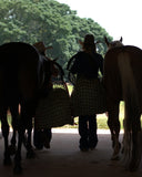 Two people leading horses out of a stable carrying hay nets with trees in the background