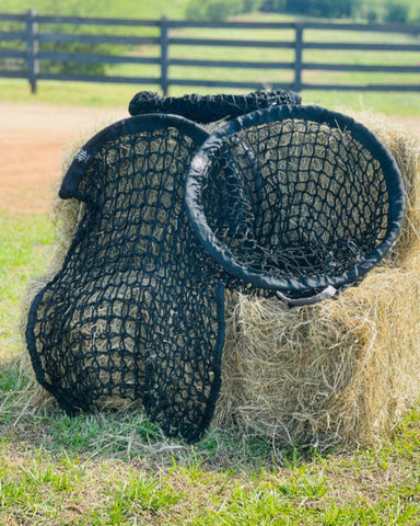 Black netted hay feeder on a stack of hay bales in an outdoor setting.