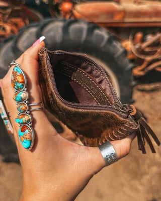 Hand holding a brown leather wallet with tassels against a rustic background with old tires and machinery.