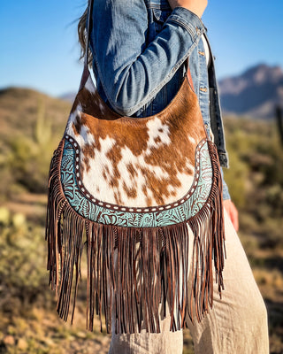 A person outdoors wearing a blue denim jacket and beige pants, carrying a large fringe shoulder bag made from brown and white cowhide with turquoise embroidered trim.